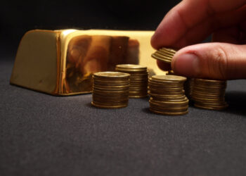 A hand stacks coins into piles on a black surface, with a gleaming gold bar in the background, hinting at the fluctuating price of gold. | GoldZeus.com