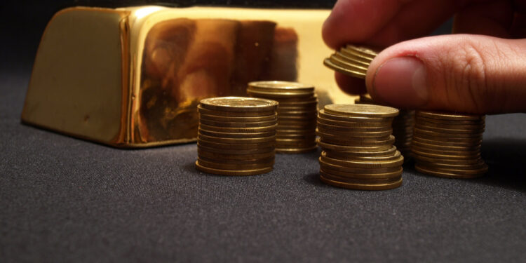 A hand stacks coins into piles on a black surface, with a gleaming gold bar in the background, hinting at the fluctuating price of gold. | GoldZeus.com