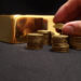A hand stacks coins into piles on a black surface, with a gleaming gold bar in the background, hinting at the fluctuating price of gold. | GoldZeus.com