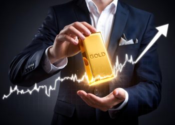 A person in a suit holds a large gold bar labeled "GOLD," illuminated by a bright, glowing light. A white, upward-trending graph line runs across the image, visually answering the question: Why Invest in Gold? It symbolizes financial growth and investment success. | GoldZeus.com