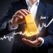 A person in a suit holds a large gold bar labeled "GOLD," illuminated by a bright, glowing light. A white, upward-trending graph line runs across the image, visually answering the question: Why Invest in Gold? It symbolizes financial growth and investment success. | GoldZeus.com