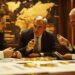 Three men in suits sit around a table covered with gold coins and documents, engaged in a serious discussion in a dimly lit office with a large world map on the wall behind them. | GoldZeus.com