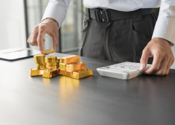 A person stands at a desk, stacking gold coins and gold bars with one hand while using a white calculator with the other hand. The scene suggests financial planning or investment analysis. | GoldZeus.com