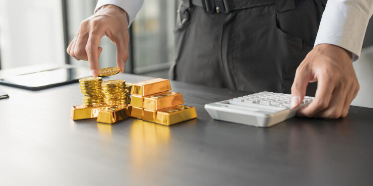 A person stands at a desk, stacking gold coins and gold bars with one hand while using a white calculator with the other hand. The scene suggests financial planning or investment analysis. | GoldZeus.com