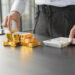 A person stands at a desk, stacking gold coins and gold bars with one hand while using a white calculator with the other hand. The scene suggests financial planning or investment analysis. | GoldZeus.com