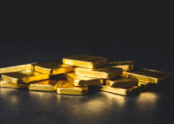 A pile of shiny gold bars stacked unevenly on a dark surface, with a black background. | GoldZeus.com