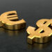 Two large, gold 3D symbols for the euro (€) and the US dollar ($) lie on a dark, textured surface, positioned side by side. | GoldZeus.com