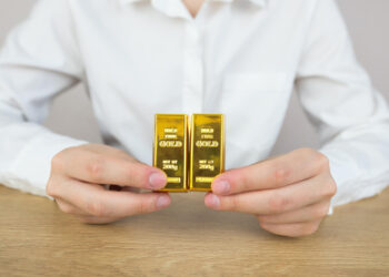 A person in a white shirt holds two gold bars side by side on a wooden table, with only their hands and torso visible. | GoldZeus.com