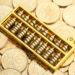 A golden abacus rests on a pile of Chinese coins with floral designs and Chinese characters. | GoldZeus.com