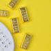 Five gold bars and a white calculator on a bright yellow background, suggesting financial calculations or investments related to gold. | GoldZeus.com