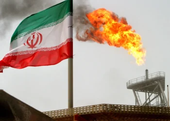 An Iranian flag waves near a large flame burning from an oil refinery tower, with smoke rising in the background on an overcast day. | GoldZeus.com