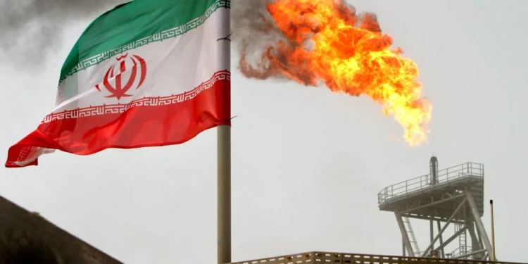 An Iranian flag waves near a large flame burning from an oil refinery tower, with smoke rising in the background on an overcast day. | GoldZeus.com