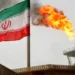 An Iranian flag waves near a large flame burning from an oil refinery tower, with smoke rising in the background on an overcast day. | GoldZeus.com